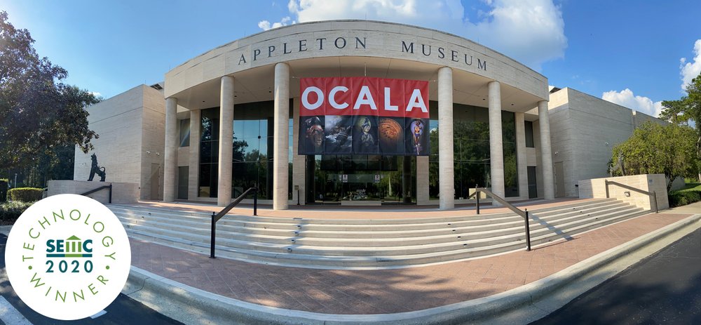 View of museum's front entrance on a sunny day. There is also a banner across the building entrance. The banner says, Ocala.