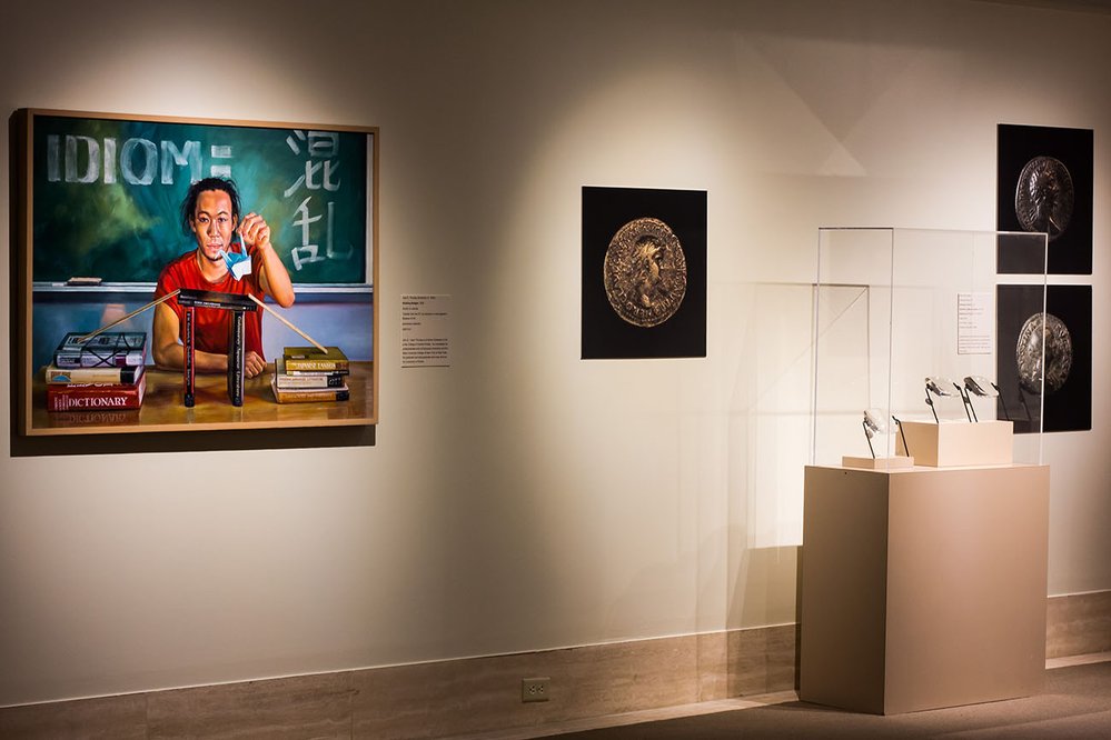 View of museum gallery. A painting of a young student is hanging on the wall, alongside enlarged posters of ancient coins.
